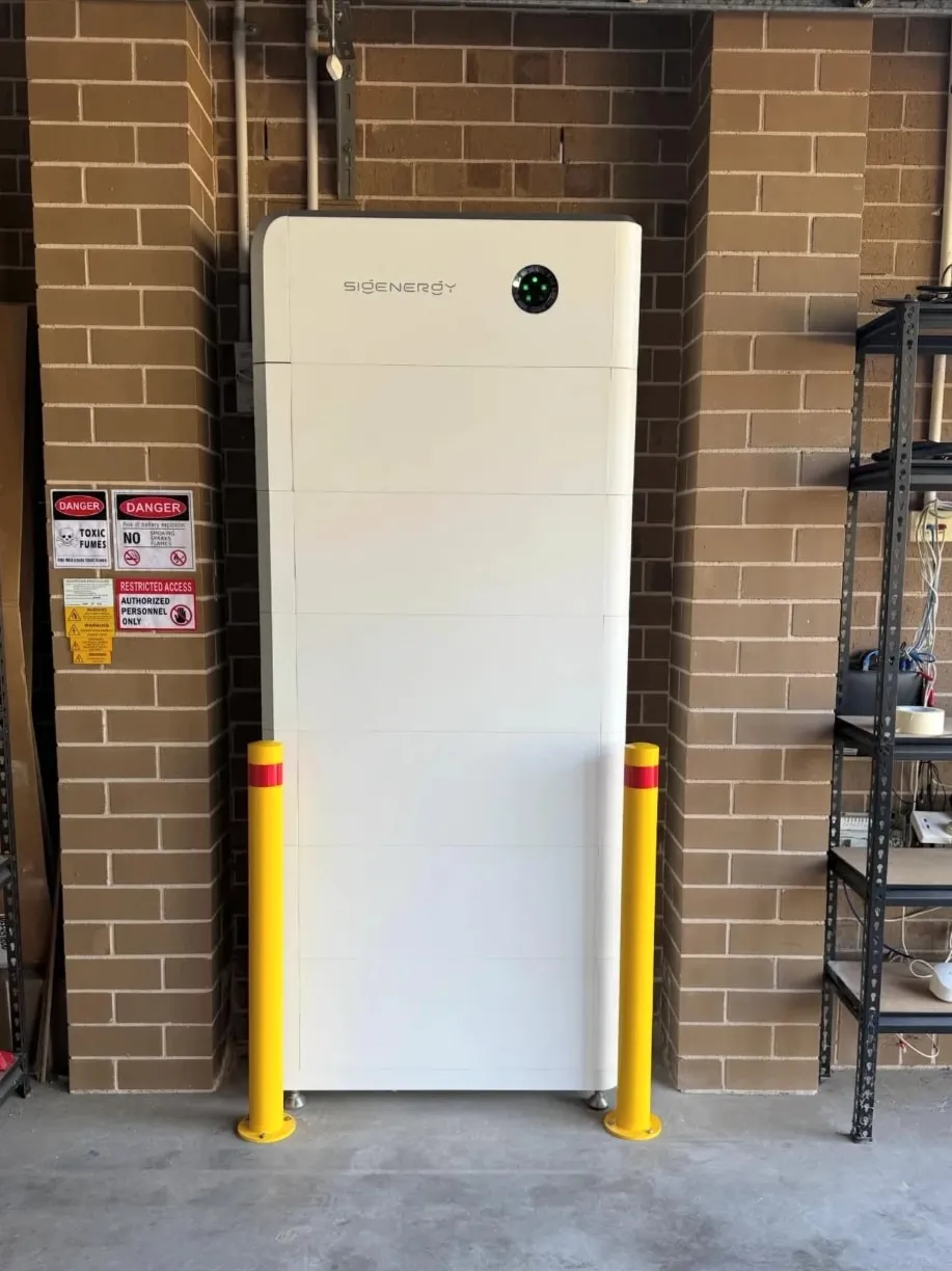 A full-height Sigenergy SigenStor stack against a brick garage wall, between two yellow bollards