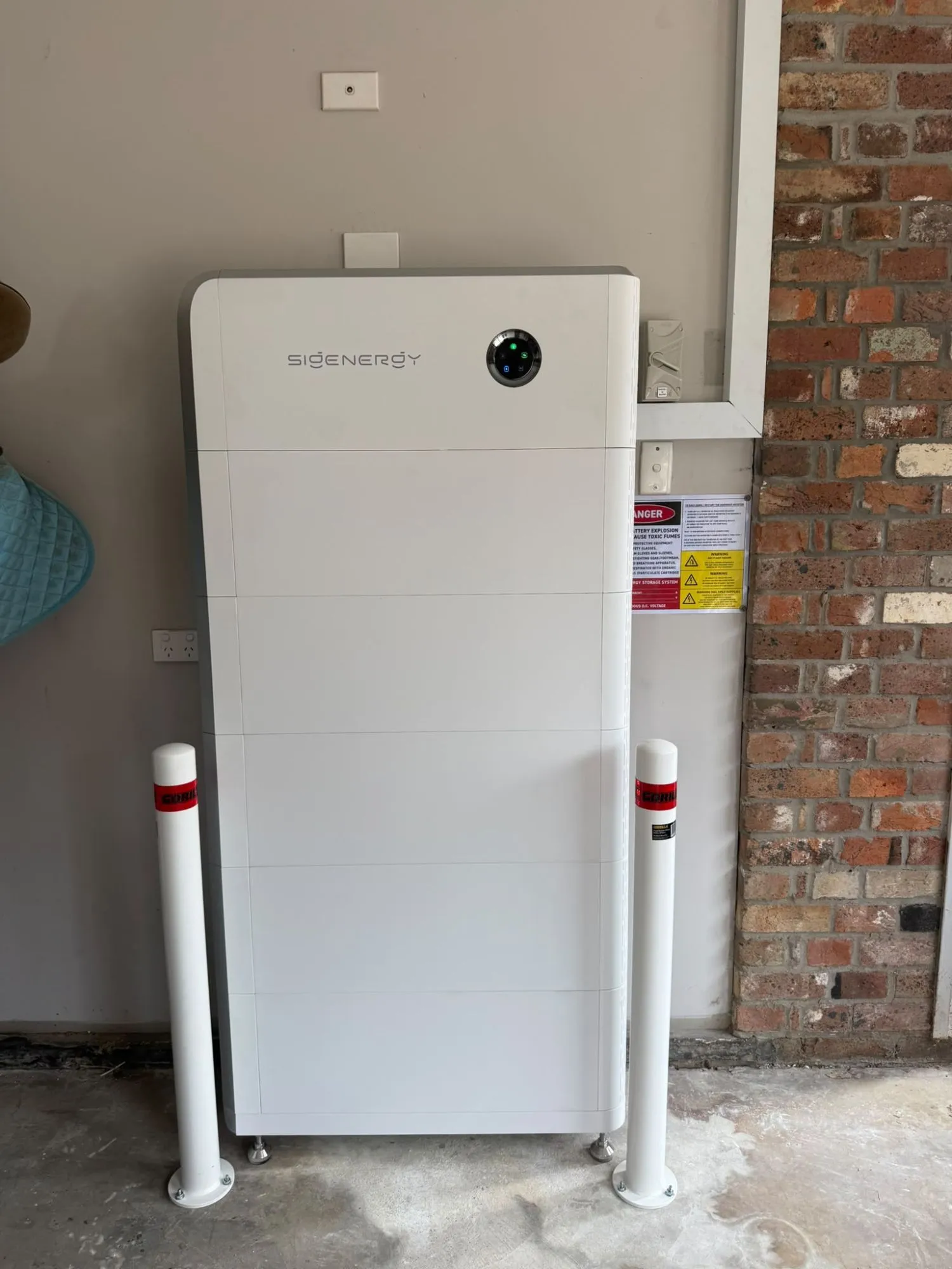 A tall white Sigenergy SigenStor stack with five battery modules under the Energy Controller, on a garage floor between two white bollards