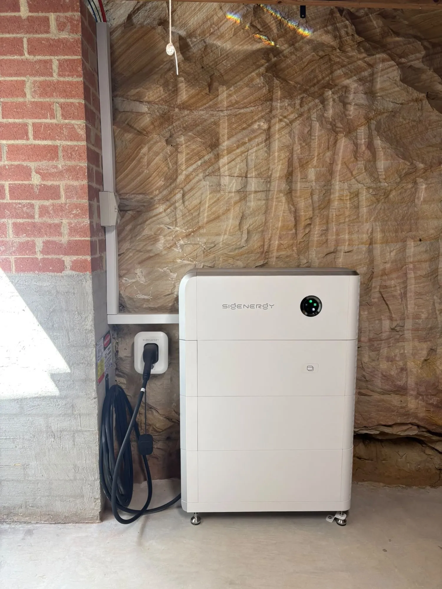 A Sigenergy SigenStor with an EV DC charging module and two battery modules against a sandstone wall, with the charging cable on a Sigenergy wall holster beside it