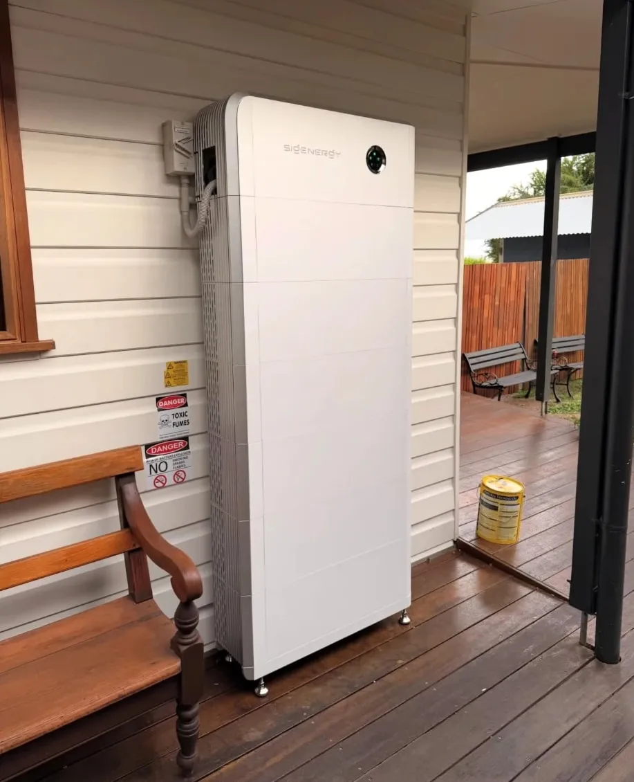 A tall Sigenergy SigenStor stack on a timber verandah against white weatherboards, beside a wooden bench