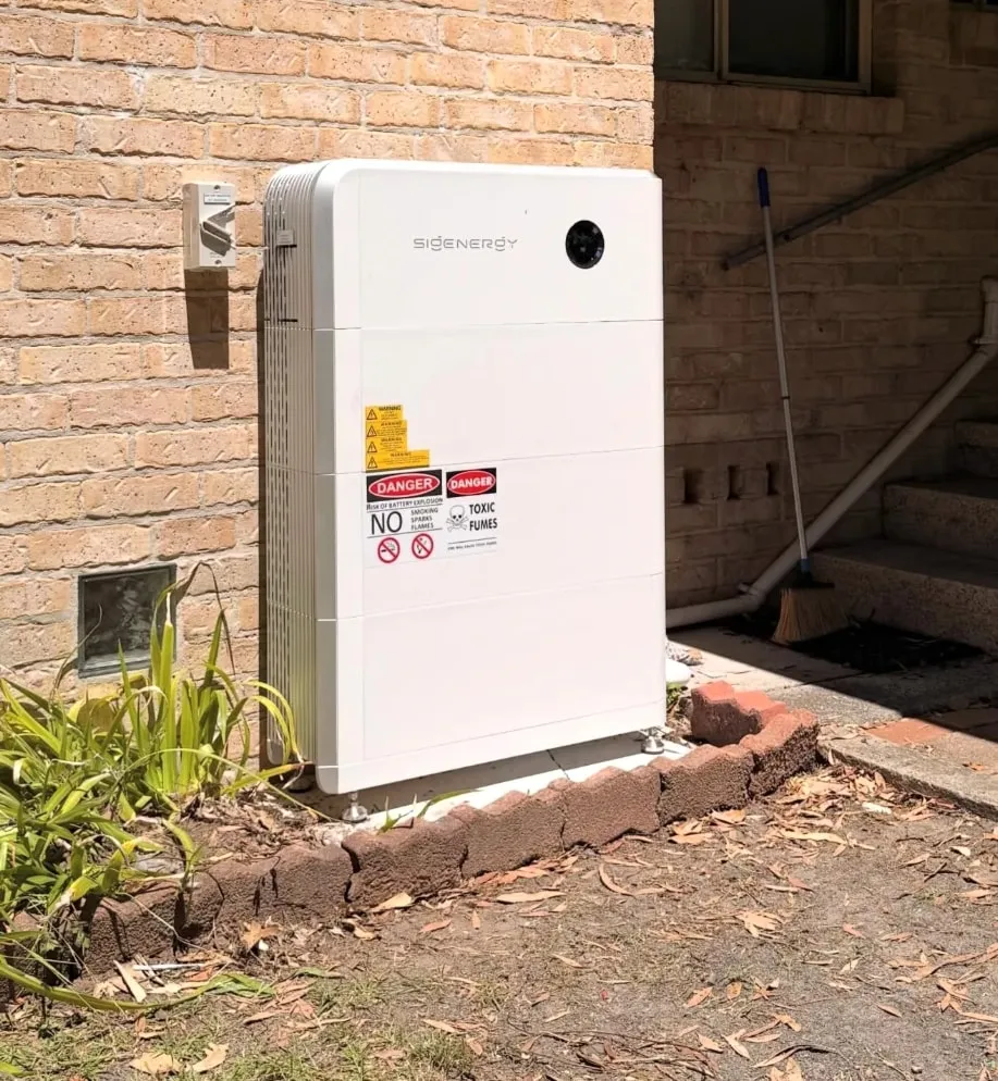 A Sigenergy SigenStor standing outside against a brick wall on its own pad, with safety signs on the front