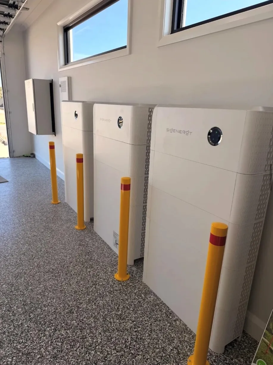 Three white Sigenergy SigenStor stacks side by side along a garage wall, each with a yellow bollard in front