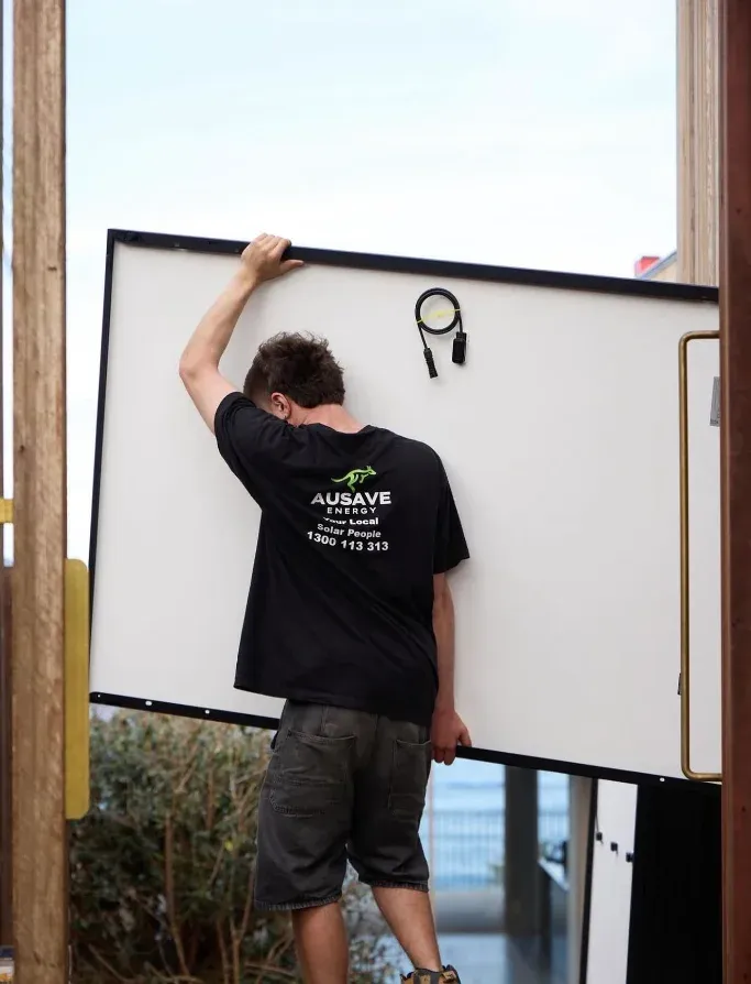 An Ausave installer carrying a solar panel, 'Your Local Solar People' printed on the back of the shirt
